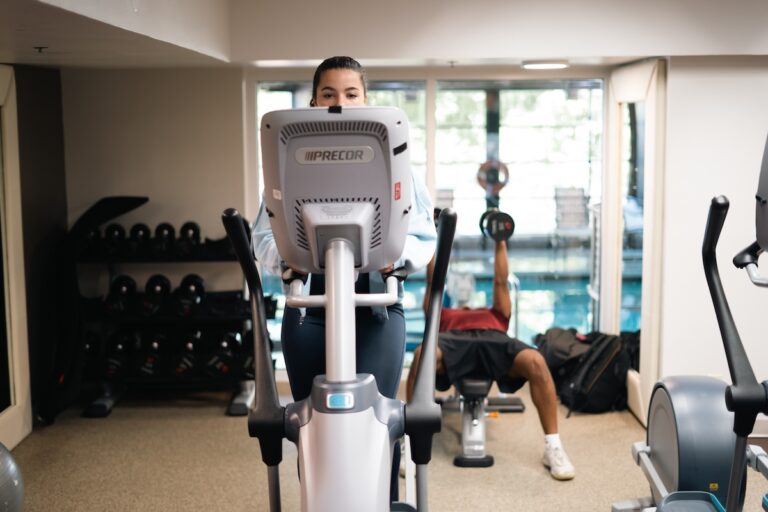 A woman works out on an elliptical machine. Another gym goer is doing chest presses with dumbbells in the background.