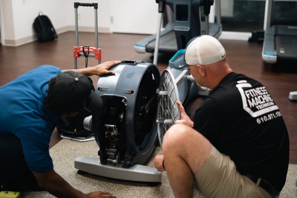 Two FMT technicians work on disassembling the back of an elliptical machine to diagnose a problem.