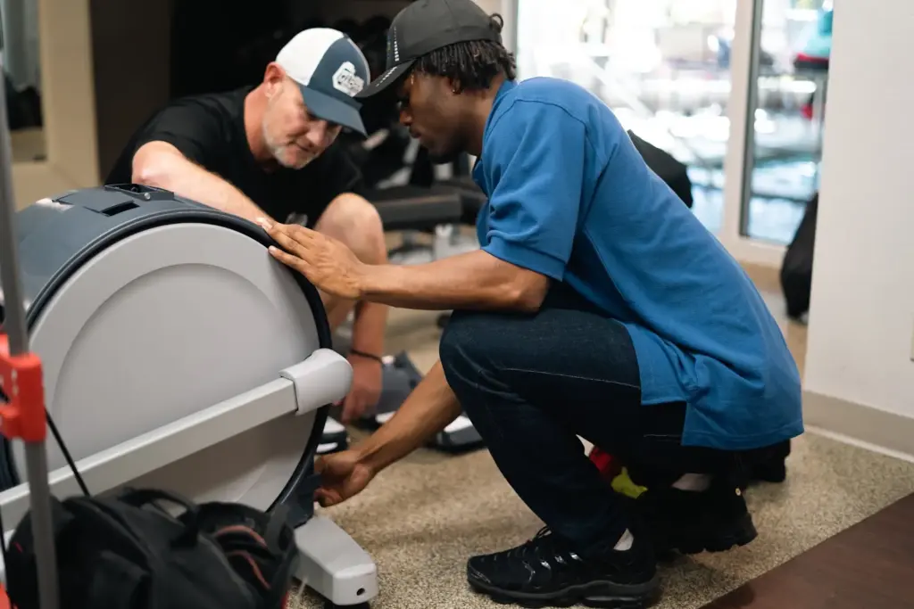 Two FMT techs work on the back of an elliptical machine. This kind of maintenance franchise is excellent because it does not require a physical footprint.